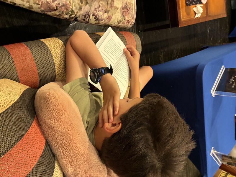 A young reader curled up on a sofa at the Bluebonnet Book Shop, most of the way through a copy of The Demon and The doG, with the signing table behind him